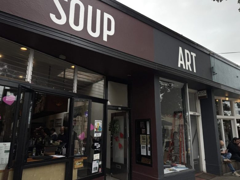 A storefront with large signs reading "SOUP" and "ART"; people are seated inside and outside the café-style establishment.