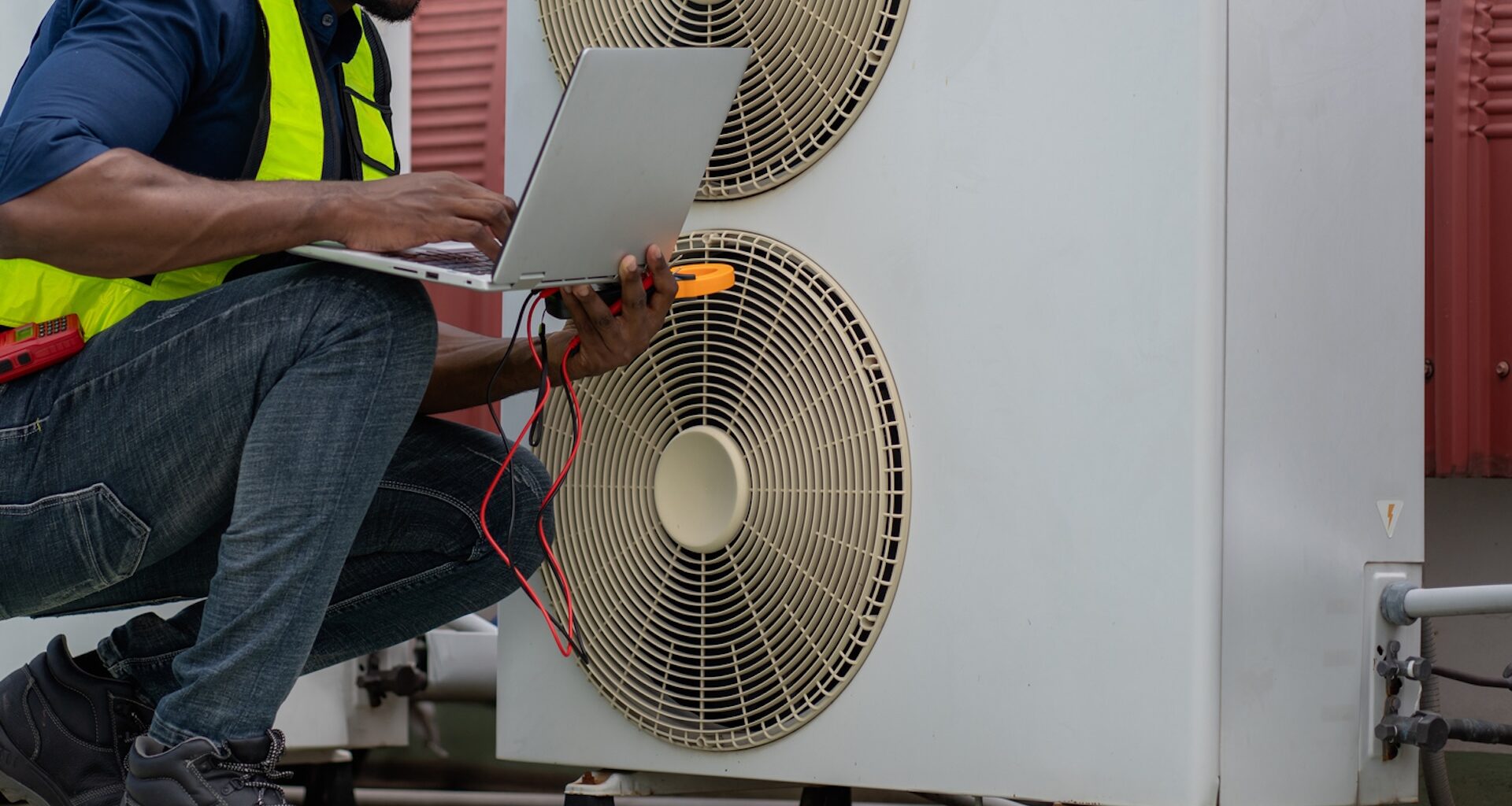 A technician in a safety vest uses a laptop to inspect a heat pump with visible electrical tools.
