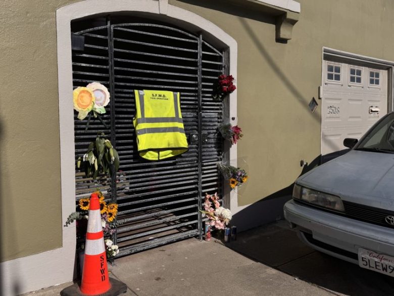A metal gate adorned with flowers, a high-visibility vest, and candles; a traffic cone and parked car are nearby on a sunlit sidewalk.