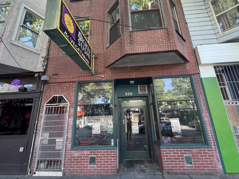 Street-level view of a red brick restaurant with a yellow and green sign reading "Sushi Vegan," located at 538, with reflections of trees in the windows.