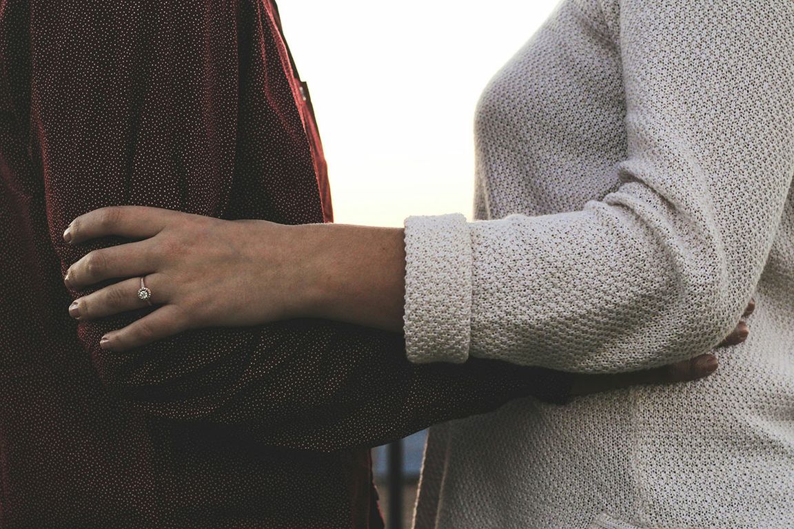 Close-up of two people embracing; one wears a round diamond solitaire ring on their left ring finger. One wears a dark burgundy dotted top, the other a light gray knit sweater.
