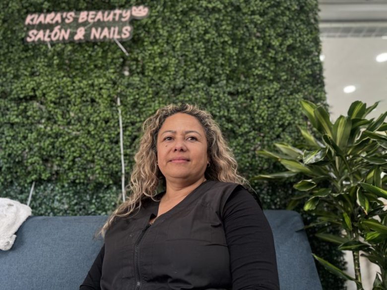 A woman with curly hair sits on a blue bench in front of a green leafy wall with a sign that reads "Kiara's Beauty Salon & Nails.