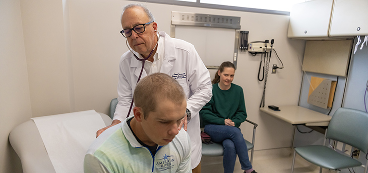 A physician examines a man, using a stethoscope on his back.