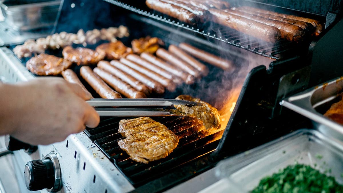 A person uses tongs to flip a piece of chicken on a grill filled with meats.