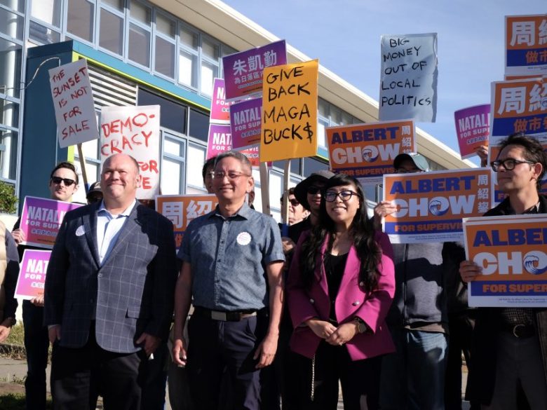 A group of people hold campaign signs for Albert Chow and Natalie Gee, with one person holding a "Give Back MAGA Bucks" sign at an outdoor political event.