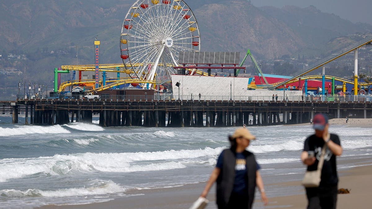 A couple walks on the beach at Santa Monica Pier with a ferris wheel in the background.