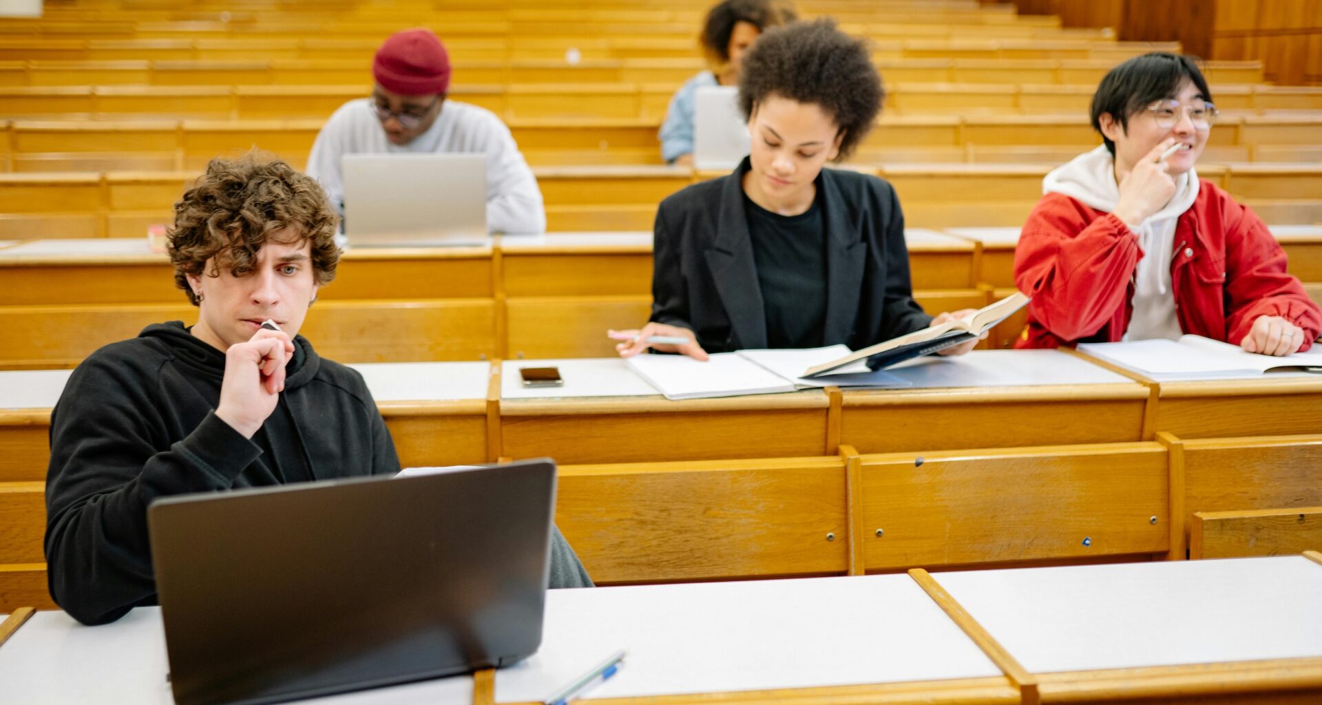 College students sit in rows at wooden tables looking at. notebooks and computer screens. A man in the front left is contemplating his laptop screen.
