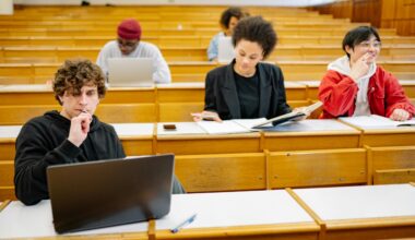 College students sit in rows at wooden tables looking at. notebooks and computer screens. A man in the front left is contemplating his laptop screen.