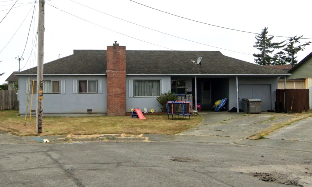 A gray house with a brick chimney and a brown roof, with a front yard containing a small red slide and a blue trampoline.