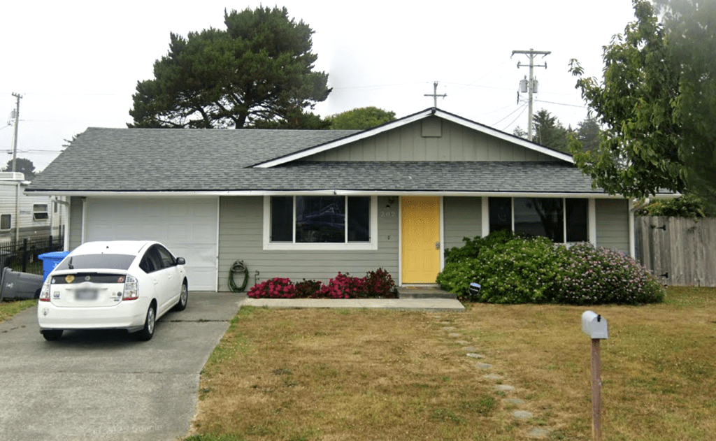 A single-story gray house with a yellow front door, a white car in the driveway, and a parched lawn.