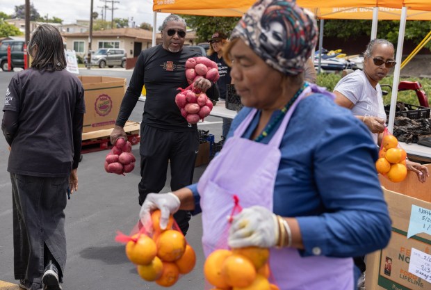 Lena Swindle, Yndia Shaw, Charles Allen and Juanita Winston, left to right, carry oranges and potatoes to a car while volunteering as people line up for food at North Park Apostolic Church on Thursday, May 28, 2026, in Lemon Grove. (Luke Johnson / The San Diego Union-Tribune)