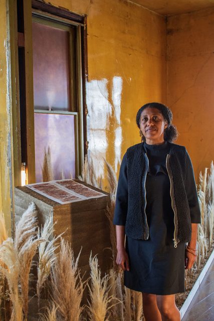 A woman stands indoors next to a window and a pedestal, surrounded by tall ornamental grasses, in a room with yellow walls.