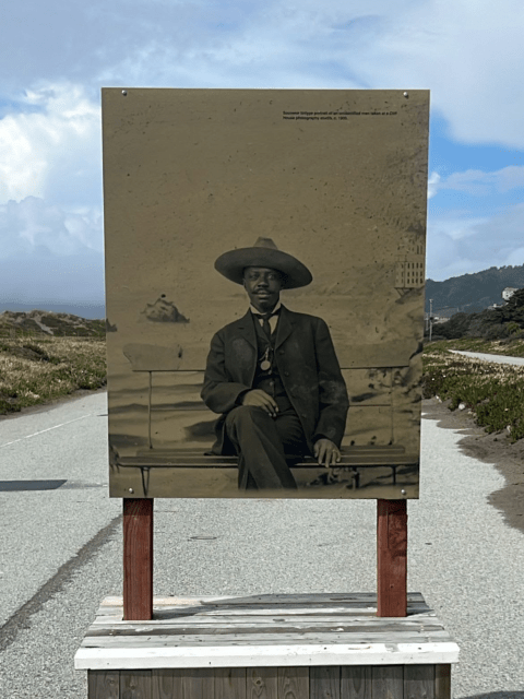 A sepia-toned historical photograph of a man in a suit and wide-brimmed hat sitting on a bench, displayed on an outdoor sign along a paved path.
