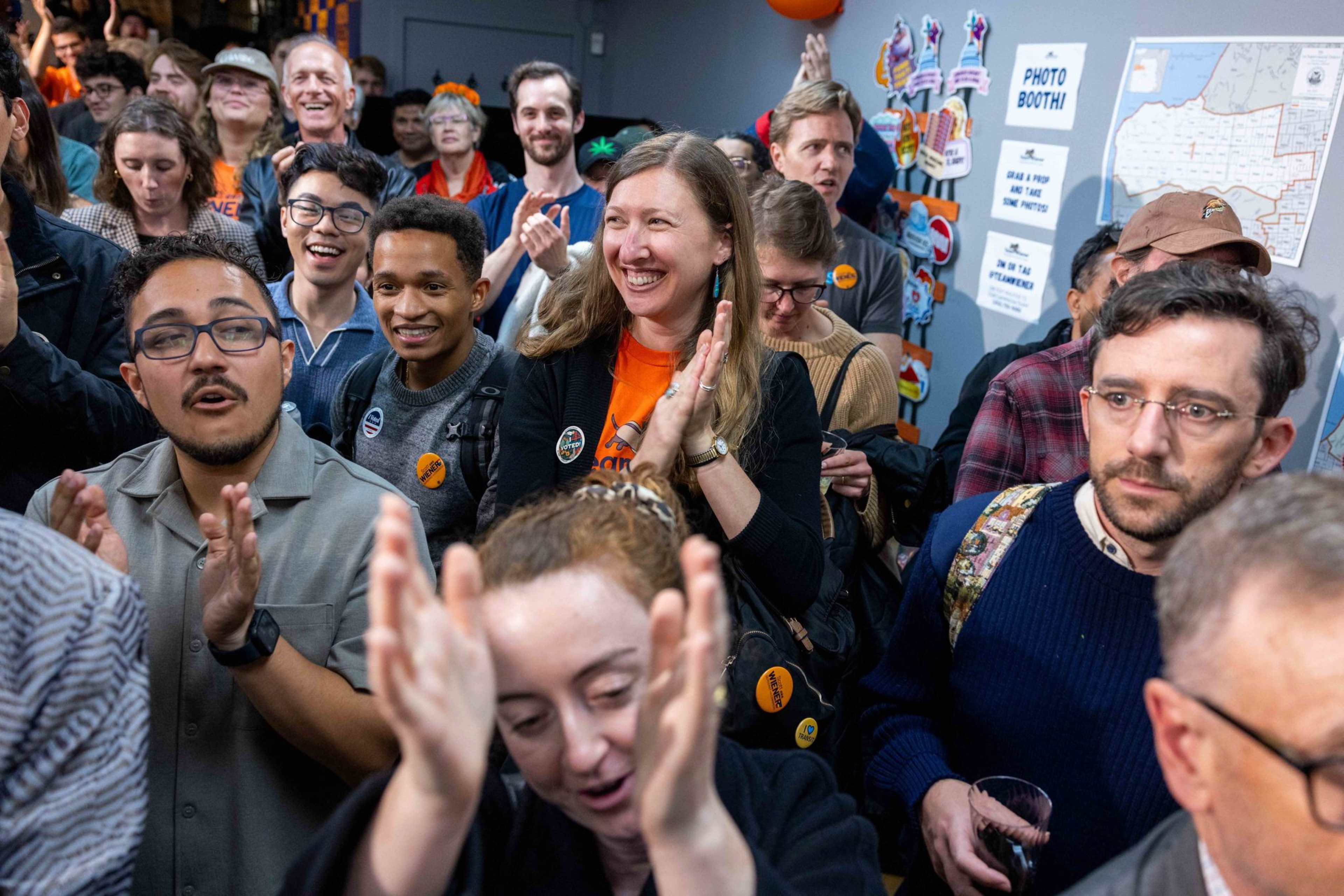 A diverse group of people are gathered closely, clapping and smiling, with some wearing buttons, appearing joyful and engaged in an indoor event.