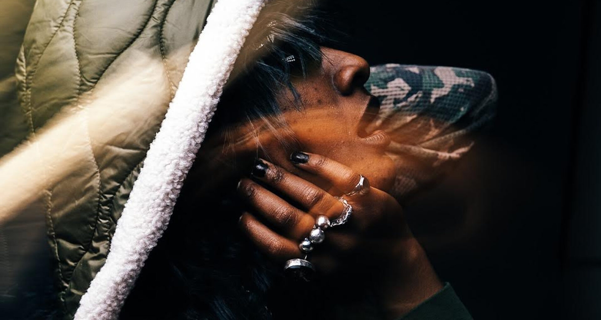 A Black face framed by a hand with multiple rings and a puffy hoddie, looking upward and to the right