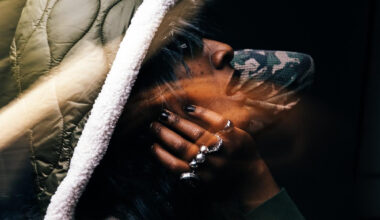 A Black face framed by a hand with multiple rings and a puffy hoddie, looking upward and to the right
