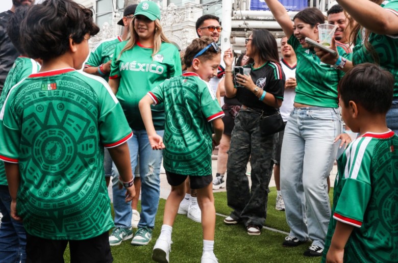 A little boy with sunglasses on his head ops with one arm in front of him and one arm behind as he stands amidst a circle of people, most of them in green jerseys. One T-shirt reads "Mexico" on the front.