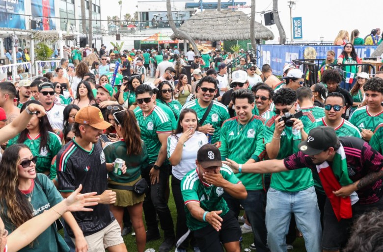 A beach hut can be seen behind dozens of people, most wearing green jerseys. In the foreground, a man points with his flat hand and covers his mouth while others pat him on the back or take photos. A banner in the background on a lamp post reads "Belmont Park."