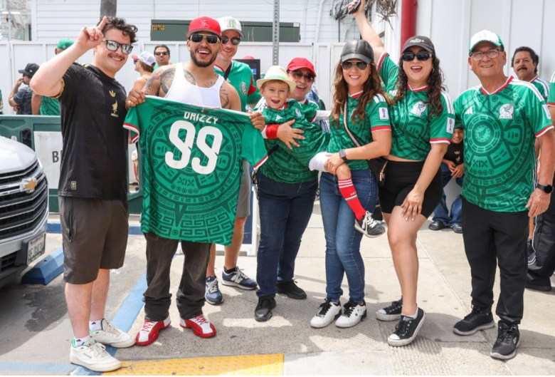 Eight people, a couple of them older, several in their 30s or 40s stand in a line smiling. Most of them are wearing green jerseys and sunglasses. Two women in the center hold a smiling little boy in a hat.