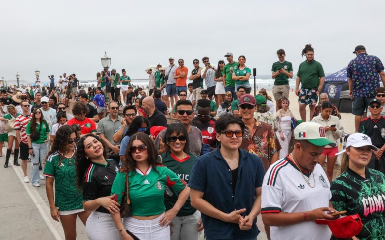 Dozens of people, many of them dressed in green, and some wearing baseball caps, line an esplanade in front of beach with the ocean visible in the background. Some stand on a wall that divides the walkway from the beach.