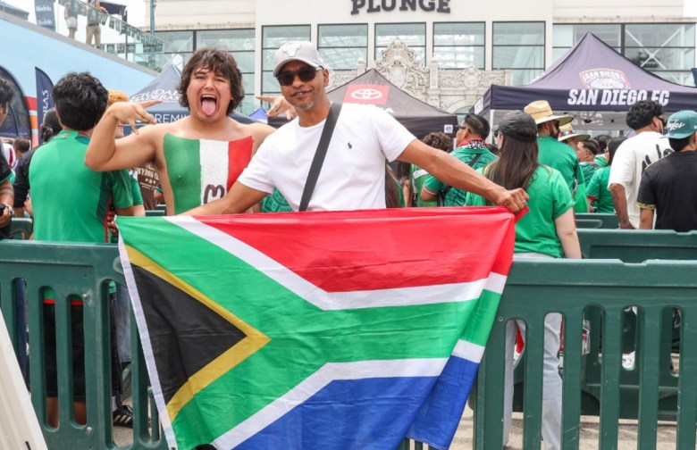 A man holds South Africa's flag, red, green and blue, while another man has the Mexican flag's green, white and red painted on his chest. They are standing behind barriers in front of a building that has a sign on it, "Plunge" and canopies that show "San Diego FC" and a symbol for Toyota.