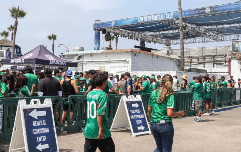 The structure behind a stage looms above many people dressed in green jerseys standing in line behind barriers in front of a canopy that reads "San Diego FC." Palm trees can be seen in the background.