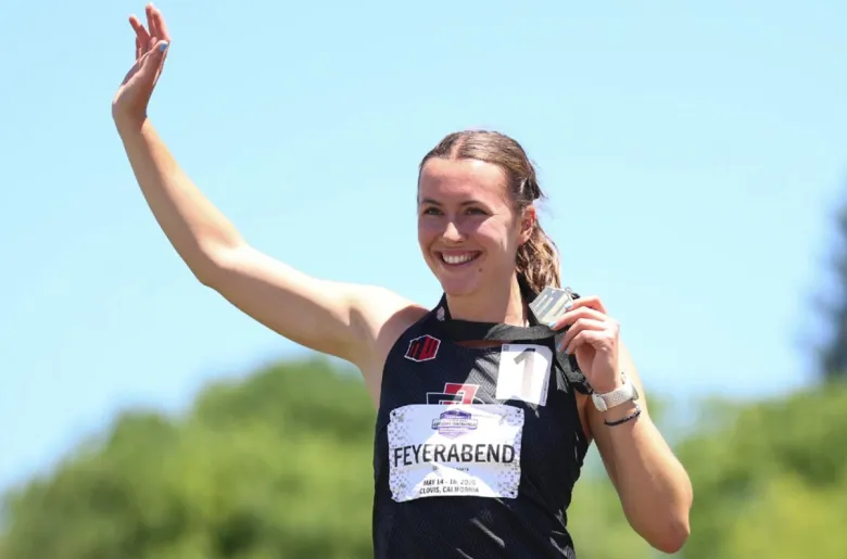 A woman waves with one arm while holding up a medal with her left hand. Her bib reads "Feyerabend." 
