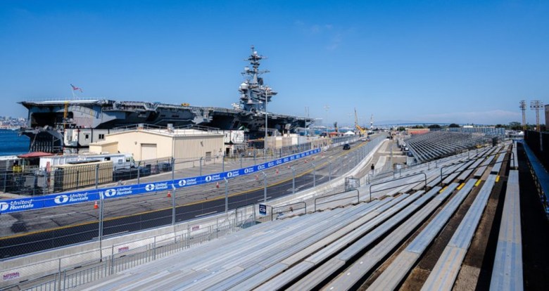 Grandstands have the view of a long track with the deck of large military ship in the background.