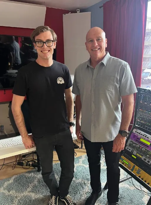 Two men stand side by side, the one on the right bald and older than the one on the left, who wears glasses.