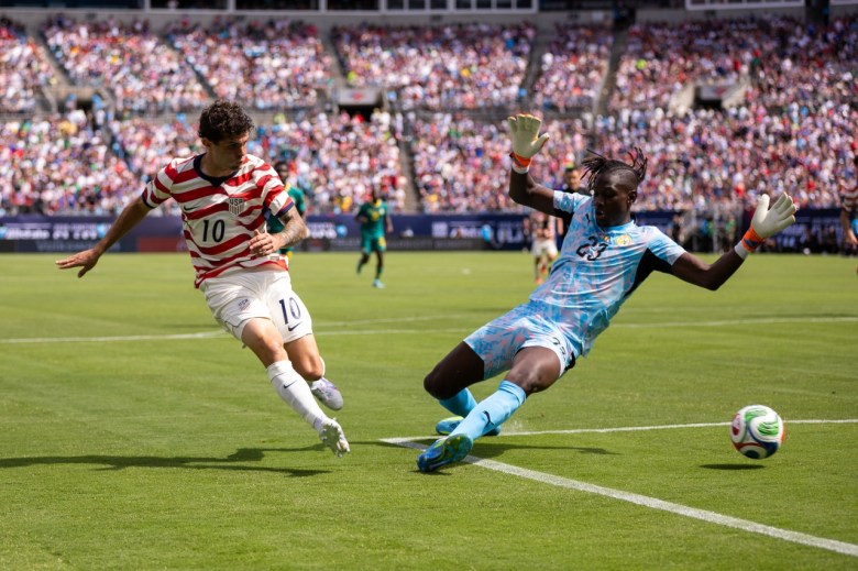 A man runs as an opponent slides, with the ball eluding him, on the soccer pitch with a packed crowd visible in the stands.