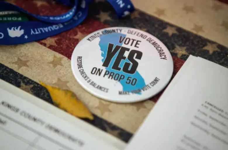 A button reads “Vote Yes on Prop 50” set against a backdrop of red, white and blue cloth with starts on it.