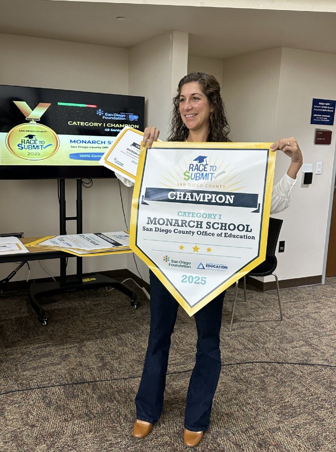 A woman in an office space holds up a banner that reads, "Race to Submit: San Diego County champion."