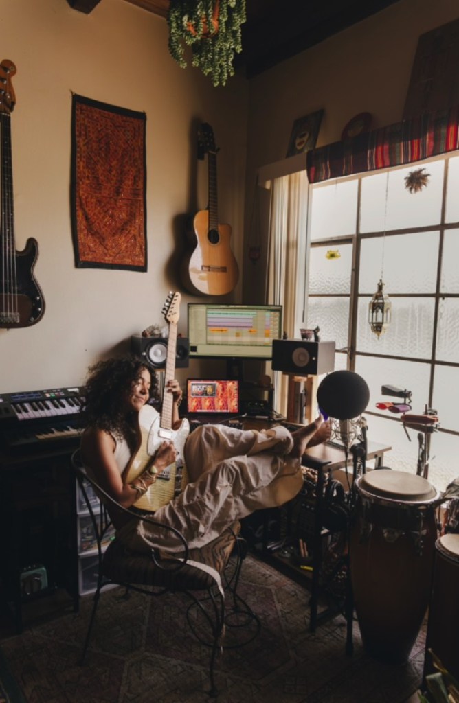 A woman sits in a home studio in front of a picture window holding a guitar pointed in the air. There is art on the wall and drums, keyboards and other instruments in the room.