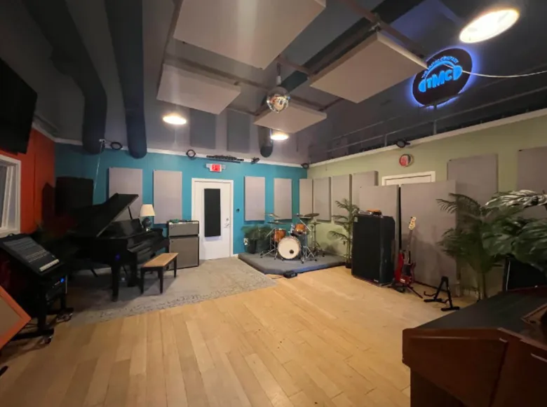 A large room with a hardwood floor. A piano sits on a rug in one corner and drum set in the opposite corner. An amplifier is on the side of the room with a guitar on a stand.