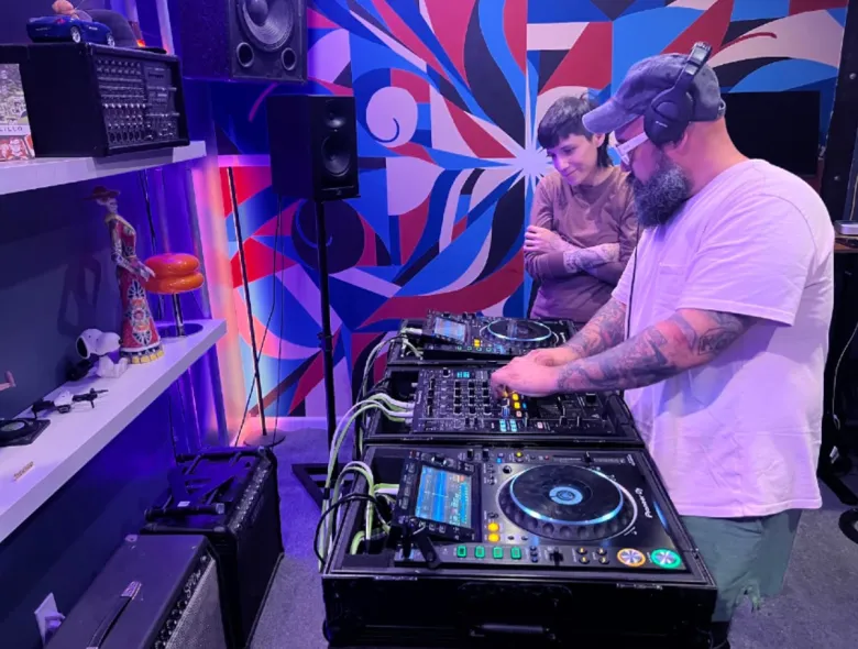 Two men watch as one works on a mixer with turntables in a recording studio. Large speakers line a back wall.