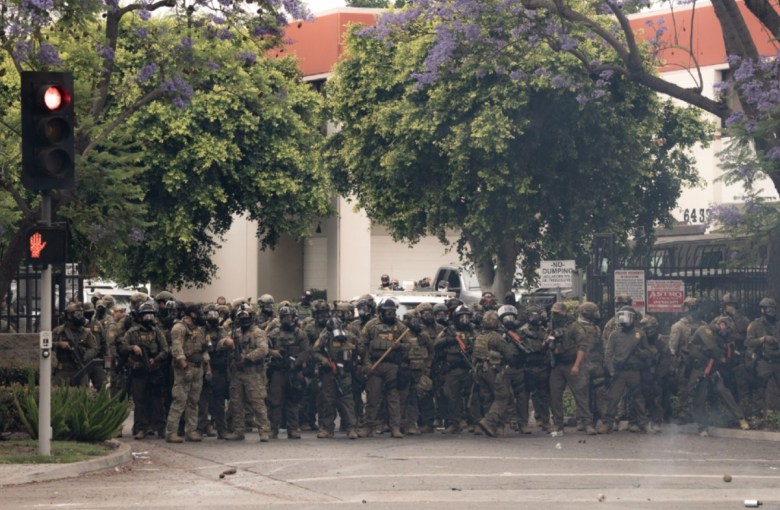 A crowd of people dressed in fatigues wearing gear, including masks and plastic face shields, and some bearing arms, stand in front of a building with a open fence visible on both sides.