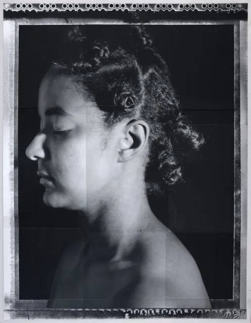 A black and white portrait of a person with closed eyes in profile view, showing short curled hair against a dark background.