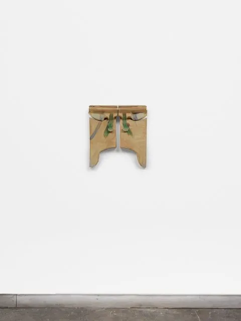 A small, abstract wooden sculpture with green accents is mounted on a white wall above a concrete floor.