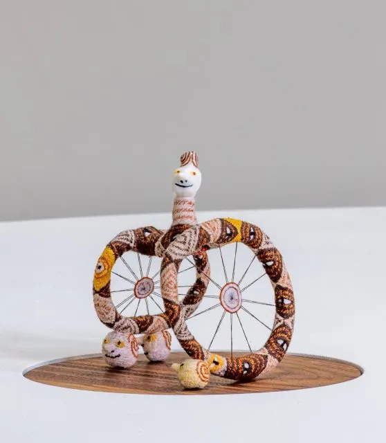 A small whimsical sculpture featuring two large wheel-like shapes and a smiling, snake-like figure with a white face, displayed on a round wooden base against a plain background.