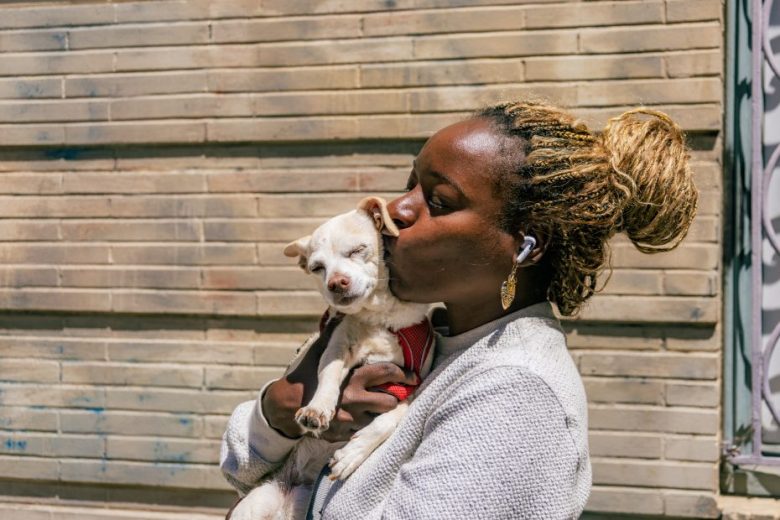 A woman with braided hair kisses a small white dog while holding it in her arms, standing in front of a beige brick wall.