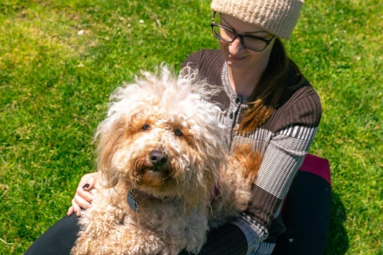 A woman wearing glasses and a knit hat sits on grass with a curly-haired dog on her lap, both looking calm and relaxed.