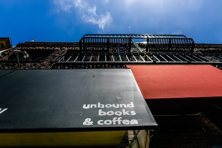 A low-angle view of a brick building with black and red awnings; the black awning reads “unbound books & coffee” in white lowercase letters.