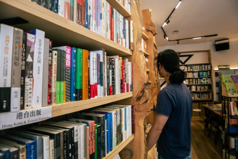 A person with long hair browses bookshelves filled with books, many with Chinese titles, in a well-lit bookstore.