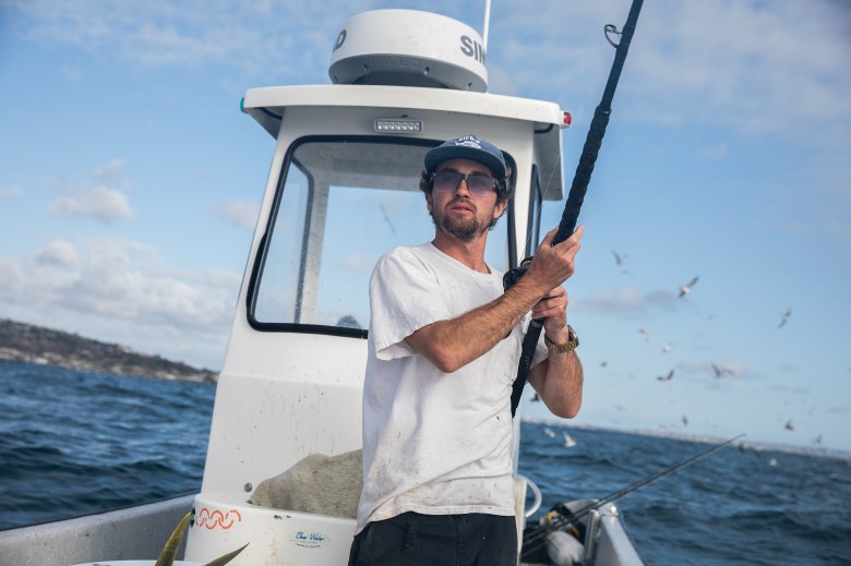 A person looks off into the distance as they hold a fishing rod while standing in the middle of a small white fishing boat that sways among ocean waves during a sunny afternoon. The person wears a hat, sunglasses and a white shirt.