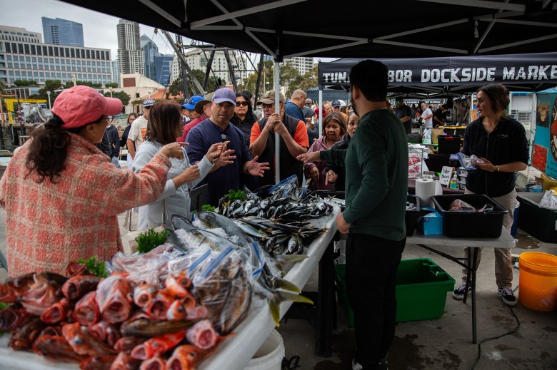 A group of people gesture and talk to one another as they stand in front of a booth at a fish market.