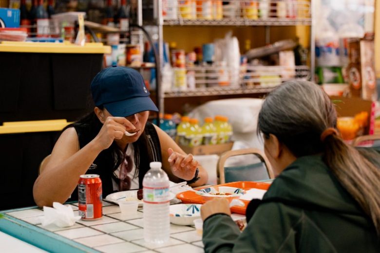 Two people sit at a table eating a meal in a store or café, with food trays, a can of soda, and a water bottle visible on the tiled tabletop. Shelves of goods are in the background.