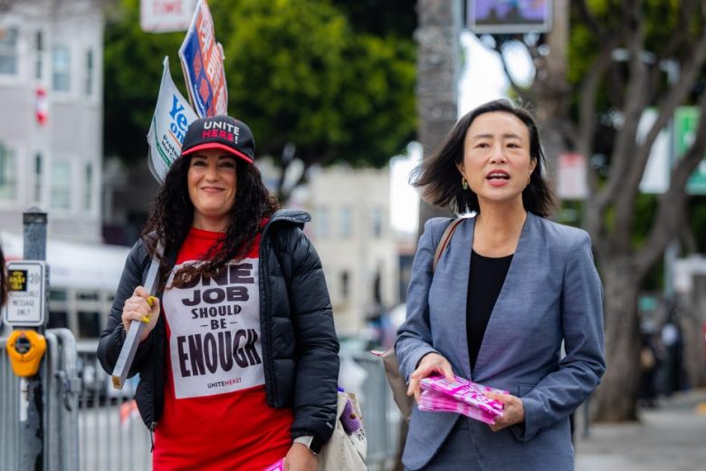 Two women stand outdoors; one holds protest signs and wears a "One Job Should Be Enough" shirt, while the other holds pamphlets and wears a gray blazer.