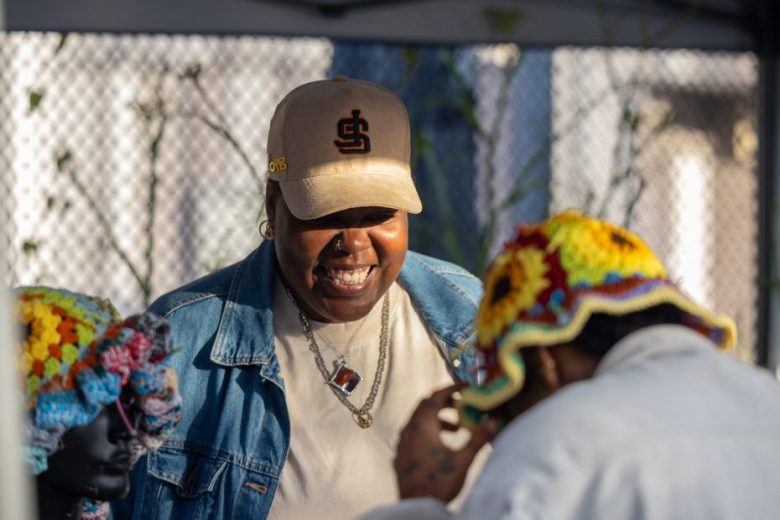 A person wearing a beige cap and denim jacket smiles while standing next to another person in a crocheted hat at an outdoor market booth.