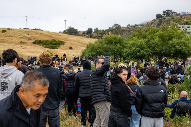 A large group of people, many in jackets, gather outdoors in a grassy area near trees and hills under a cloudy sky.