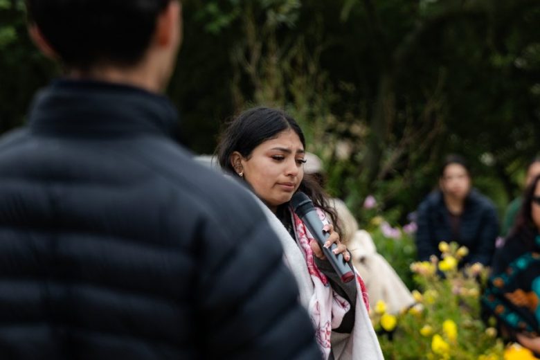 A woman holds a microphone and speaks outdoors among flowers, with several people listening in the background.